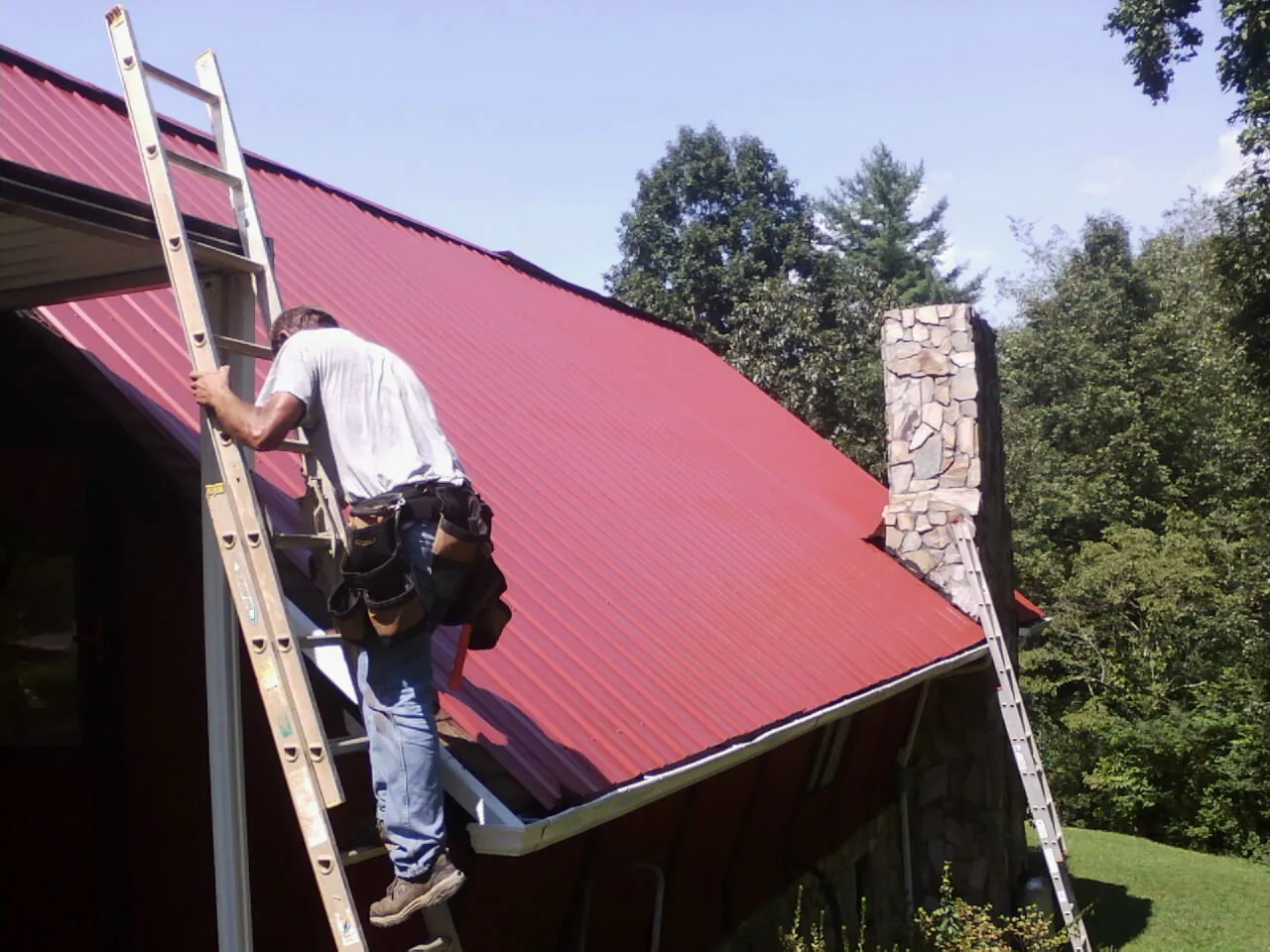 Roofer on a ladder beside a red metal roof and stone chimney — Soffit & Fascia Repair by Roofing Brothers in Sebeka